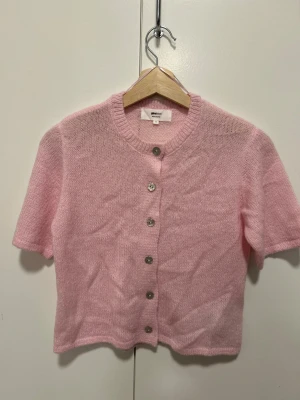 Rosa stickad kofta från Gina Tricot - Supergullig ljusrosa stickad kofta från Gina Tricot med korta ärmar och pärlemorknappar framtill. Mjuk och fluffig känsla, perfekt för vår och sommar. Rund halsringning och normal passform, enkel att styla till jeans eller kjol.