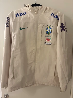 Beige Nike Brasil vindjacka med huva - Snygg beige vindjacka från Nike med Brasil-logga och sponsormärken som Itaú och Vivo. Jackan har huva, hel dragkedja framtill och lång ärm. Materialet är lätt och syntetiskt, perfekt för dig som gillar sportig stil och fotboll. Använd 1 gång