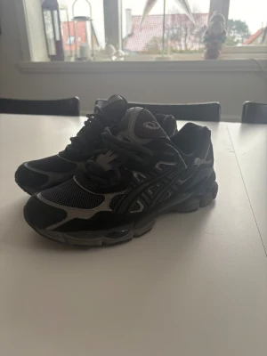 Svarta Asics Gel Nyc sneakers med mesh och skinn - Säljer ett par svarta Asics sneakers med grå detaljer. Skorna har en chunky sula, mesh-överdel för bra ventilation och inslag av syntet och skinn. Rund tå och snörning framtill. Perfekt för dig som gillar sportig och modern stil.