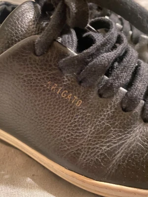 Svarta sneakers från Axel Arigato - Snygga svarta sneakers från Axel Arigato i äkta skinn med vit sula. Klassisk låg modell med rund tå och svarta snören. Diskret guldtext på sidan och stilren design som passar till det mesta. Perfekta för dig som gillar minimalistisk och clean stil.🩷