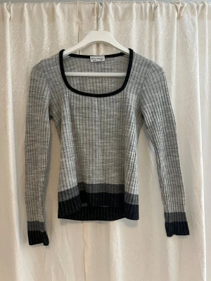 Grå ribbad långärmad tröja från Van Studio - Snygg grå ribbad tröja med svarta detaljer vid halsringning, nederkant och ärmslut. Tröjan har lång ärm och en bred, rundad hals. Perfekt för dig som gillar stilrena och enkla plagg med lite edge.