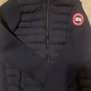 Mörkblå pufferjacka från Canada Goose - Snygg mörkblå pufferjacka från Canada Goose med quiltad framsida och dragkedja. Jackan har långärmade släta ärmar och klassisk logga på vänster arm. Perfekt för kyliga dagar och har en stilren look som funkar till det mesta.