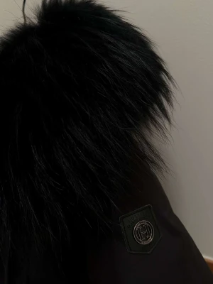 Svart vinterjacka Hollies med päls - Ordinarie pris: 5249kr Snygg svart vinterjacka från Hollies med stor fluffig päls runt huvan. Jackan har flera fickor, dragkedja och knappar framtill samt coola detaljer. Perfekt för kalla dagar när du vill vara både varm och stilren.