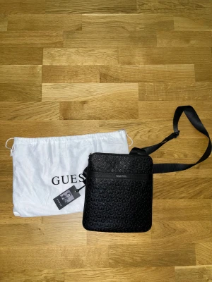 Svart axelväska från Guess - Snygg svart axelväska från Guess med justerbar rem och präglat logomönster över hela väskan. Väskan har en stilren, fyrkantig form och dragkedja upptill. Materialet är skinnimitation och den kommer med original dustbag och tag.
