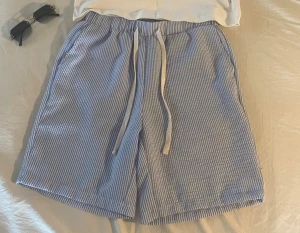 Randiga blå-vita linnen shorts med snörning - Säljer ett par chill randiga shorts i blått och vitt med elastisk midja och snörning framtill. De har en avslappnad passform, sidofickor och en bakficka. Perfekta för varma dagar och softa outfits. Materialet känns lätt och luftigt.