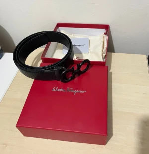 Svart skärp från Salvatore Ferragamo - Stilrent svart bälte från Salvatore Ferragamo med ikoniskt dubbel-Gancini-spänne i svart metall. Tillverkat i präglat skinn och levereras i original röd ask med logga. Perfekt accessoar för att lyfta din outfit.