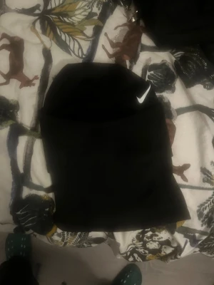 Nike Hyperwarm svart mössa - Svart Nike Hyperwarm mössa med klassisk vit swoosh-logga framtill. Tillverkad i mjukt och värmande syntetmaterial, perfekt för kalla dagar. Snygg och enkel design med diskret Nike-märke på baksidan. Passar dig som gillar sportig och clean stil.