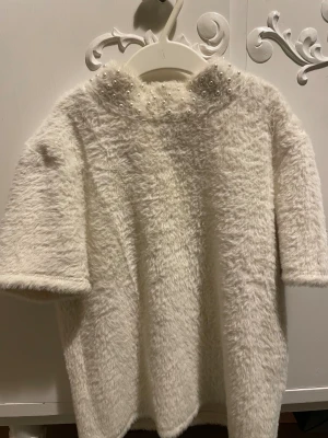 Vit fluffig tröja med pärlor från Zara - Supermjuk och fluffig vit tröja från Zara i storlek M. Tröjan har korta ärmar och är dekorerad med pärlor runt halsen för en extra cool touch. Materialet är pälsliknande syntet som känns riktigt mysigt. Perfekt för dig som gillar statement-plagg. Passar även storlek S!