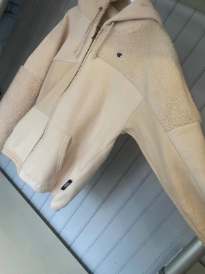 Beige teddyjacka med huva från Champion - Mysig teddyjacka från Champion i beige med huva och dragkedja. Jackan har en fluffig teddystruktur upptill och på ärmarna, samt släta partier på kroppen. Praktiska fickor framtill och snörning i huvan. Endast använd en gång. Det är storlek L står det men tycker att den sitter ganska bra på mig som har S om man vill ha den lite oversized  