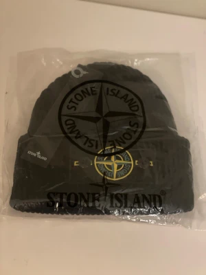 Svart mössa från Stone Island - Svart ribbstickad mössa från Stone Island med klassisk logotyp-patch framtill i gult och grönt. Mössan har uppvikt kant och är tillverkad i ett mjukt stickat material som håller dig varm och snygg under kalla dagar. Perfekt accessoar för streetwear-stilen.