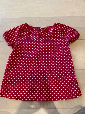 Röd blus med hjärtan från Lindex - Supersöt röd blus från Lindex med vita hjärtan över hela tyget. Blusen har korta ärmar och en liten knappdetalj vid halsen. Perfekt för dig som gillar färg och gulliga mönster!