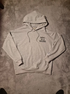 Grå hoodie från Gallery Dept. M - Snygg ljusgrå hoodie från Gallery Dept. med svart tryck 'DÉPT. de la GALERIE' på bröstet och en patch på ärmen. Tröjan har huva med snörning, känguruficka och ribbade muddar. Perfekt för dig som gillar streetwear och vill ha något chill och stilrent.