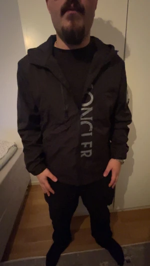 Svart vindjacka Moncler Xl/L - Svart vindjacka från Moncler i storlek Xl/L med stor logotyp framtill och klassisk Moncler-patch på ärmen. Jackan har huva, dragkedja och snörning vid halsen. Materialet är lätt och syntetiskt, perfekt för blåsiga dagar. Snygg och stilren design med diskreta detaljer. (PRIS går att diskutera)