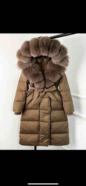 Brun lång pufferjacka med päls - Säljer en brun lång pufferjacka med stor fluffig päls runt huvan och ett snyggt bälte i midjan. Jackan har långa ärmar och quiltad design, perfekt för kalla dagar. Stängs med knytband och har en riktigt lyxig vibe.