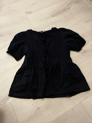 Mörkblå blus med knyt och puffärm - Säljer en mörkblå blus med korta puffärmar och volang nedtill. Blusen har två dekorativa knyt framtill och en rund halsringning. Materialet känns mjukt och lätt, perfekt för dig som gillar en söt och trendig look. Passar storlek 150-170