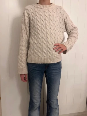 Beige kabelstickad tröja - Säljer en beige kabelstickad tröja med rund hals och långa ärmar. Tröjan har ett snyggt flätmönster över hela och breda ribbade muddar vid ärmslut och nederkant. Perfekt för dig som gillar en chill och klassisk stil, det är storlek 146/152. Och det är två tröjor 