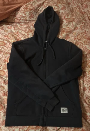 Svart hoodie från Andy by Frank Dandy - Svart hoodie med dragkedja och huva från Andy by Frank Dandy. Tröjan har snörning i huvan, två fickor fram och en liten vit logolapp på ena fickan. Materialet är 70% bomull och 30% polyester, vilket gör den mjuk och skön. Perfekt för chill dagar.