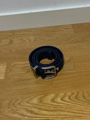 Mörkblå Levi's bälte - Snyggt mörkblått bälte från Levi's i textil med tydlig vit logga tryckt längs hela bältet. Klassisk metallspänne i silverfärg och stilren design som passar till jeans eller chinos. Perfekt accessoar för att lyfta din outfit med en sportig touch.