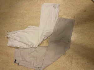 Två par linnebyxor lager 157 - Säljer två par beige byxor i bomull. Ena paret har resår och snörning i midjan för en avslappnad look, medan det andra paret har klassisk knapp och gylf. Perfekta för en chill och stilren outfit.