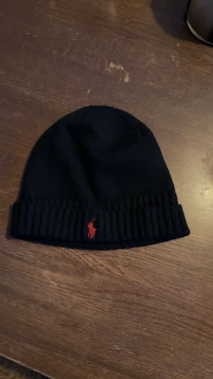 Svart stickad mössa från Polo Ralph Lauren - Svart stickad mössa med uppvikt kant och röd Polo Ralph Lauren-logga broderad framtill. Klassisk och enkel design som passar perfekt för kalla dagar. Mjuk och skön kvalitet.