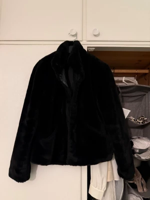 Svart pälsjacka med krage - Säljer en svart pälsjacka med mjuk och fluffig känsla. Jackan har en klassisk krage och raka, långa ärmar. Perfekt för dig som vill ha en stilren och mysig look under kyliga dagar. Enkel design utan synliga detaljer på framsidan.