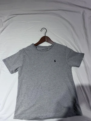 Grå t-shirt från Polo Ralph Lauren - Snygg grå t-shirt från Polo Ralph Lauren med klassisk marinblå logga broderad på bröstet. Tröjan är i nyskick förutom texten som är lite borttvättad 