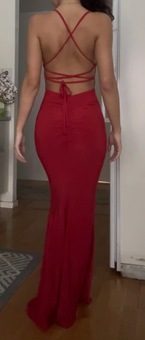 Röd långklänning med snörning - Säljer en röd långklänning med smala axelband och snygg snörning i ryggen. Klänningen har v-ringad front och figurnära passform som framhäver siluetten. Materialet är mjukt och stretchigt, perfekt för dig som vill ha en elegant look med öppen rygg och knytning.