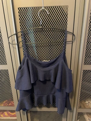 Mörkblå volangtopp från Nelly - Supersöt mörkblå linnetopp från Nelly med dubbla volanger och smala axelband. Toppen har en flowy och luftig känsla, perfekt för varma dagar. Materialet är mjukt och lätt, och volangerna ger en extra touch till outfiten.