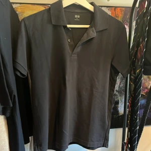 Marinblå piké T-shirt från Uniqlo - Marinblå piké t-shirt från Uniqlo med klassisk krage och två knappar vid halsen. T-shirten har korta ärmar och är gjord i mjuk bomull som känns skön mot huden. Passformen är normal och stilen är clean och enkel, perfekt till jeans eller shorts. Hör gärna av er vid funderingar eller prisförslag. 