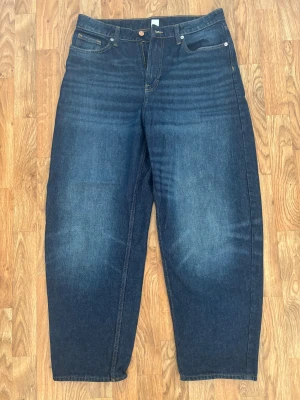 Blå barrel jeans från &Denim, strl 40 - Säljer ett par mörkblå barrel jeans från &Denim i storlek 40. Modellen har loose fit och hög midja, med klassiska fem fickor och raka ben. Jeansen är tillverkade i denim och har snygga kontrastsömmar. Perfekt för dig som gillar avslappnad stil.
