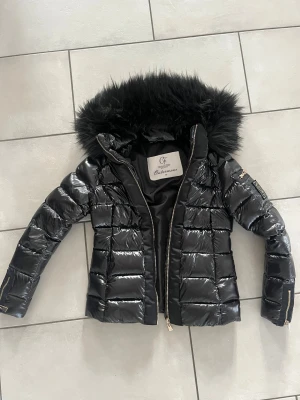 Svart pufferjacka från Chiara Forthi - Snygg svart pufferjacka från Chiara Forthi med glansig finish, guldiga dragkedjor och stor fluffig huva med päls. Perfekt för kalla dagar när du vill vara både varm och stilren. Jackan är i ett väldigt fint skick och endast använd ett fåtal gånger. 