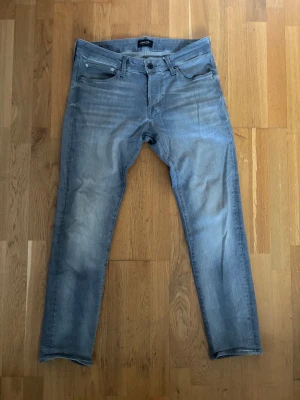 Slim Glenn ljusgrå jeans från Jack & Jones - Slim Glenn jeans från Jack & Jones i ljusgrå tvättad denim. Modellen har fem fickor, smal passform och klassisk knappgylf. Snygga sömmar på bakfickorna och diskret logotyp på linningen. Perfekt för dig som gillar en modern och stilren look.
