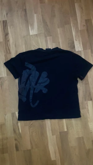 Svart t-shirt från Syna World - Svart t-shirt från Syna World i storlek L. T-shirten har ett stort, mörkgrått grafiskt tryck som går över både fram- och baksidan. Klassisk passform med rund hals och korta ärmar. Skön och mjuk känsla, perfekt till streetwear-stilen.