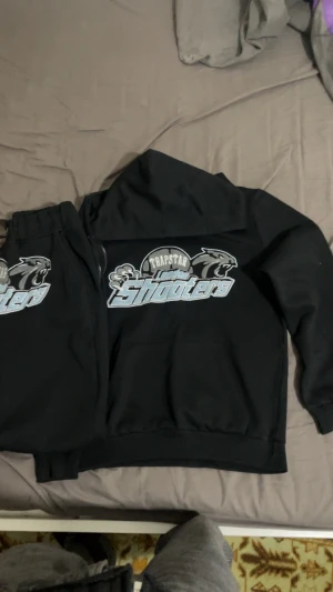 Trapstar svart traqzut med tryck - Svart hoodie från Trapstar med stort 'London Shooters' tryck i ljusblått och vitt på bröstet. Hoodien har huva och magficka, samt coolt pantermotiv och textdetaljer. Perfekt för dig som gillar streetwear och vill sticka ut med snygg grafik.