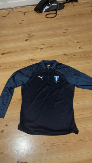 Malmö FF långärmad träningströja Puma -  OANDVÄND. Snygg långärmad fotbollströja från Puma med Malmö FF-logga på bröstet. Tröjan är mörkblå med ljusblå detaljer och har ett subtilt geometriskt mönster på axlarna. Materialet är funktionellt och andas, perfekt för träning. Halv dragkedja framtill.