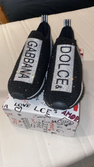 Dolce & Gabbana svarta sneakers med strass - Säljer ett par svarta Dolce & Gabbana sneakers med glittrande strassdetaljer och tydlig logga på ovansidan. Skorna har en chunky, vit och svart sula med grovt mönster och elastisk ovandel i mesh. Snygg och iögonfallande design som verkligen sticker ut.