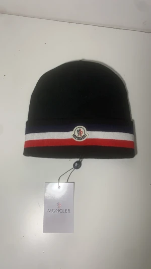 Svart Moncler mössa med ränder - Svart mössa från Moncler med bred uppvikt kant i marinblått, vitt och rött. Klassisk Moncler-logga framtill och mjukt stickat material. Säkra er mössa inför den kalla tiden och kylan.