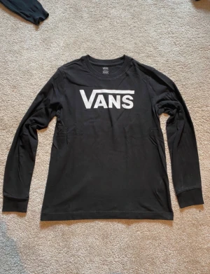 Svart långärmad Vans tröja - Svart långärmad tröja från Vans i storlek small. Tröjan har en klassisk rund hals och stort vitt Vans-tryck på bröstet. Tillverkad i mjuk bomull som är skön mot huden. Perfekt för dig som gillar streetwear och enkel stil.