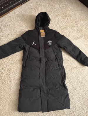 Svart lång dunjacka PSG x Jordan - Lång svart dunjacka från Nike Jordan x PSG med huva, dragkedja och två sidofickor. Jackan har PSG-logga och Jumpman-logga på bröstet. Perfekt för kalla dagar och har en clean sportig look.