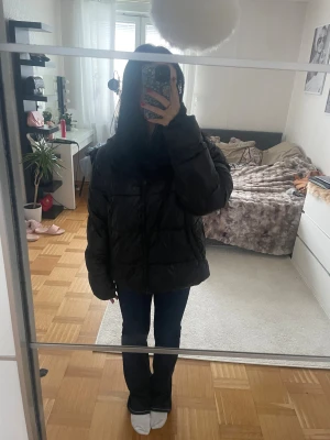 Svart pufferjacka med huva - Säljer en svart pufferjacka från lager 157 med glansig finish och huva. Jackan har dragkedja framtill och två fickor på sidorna. Perfekt för kalla dagar och har en oversized vibe. Passar dig som gillar streetstyle och vill hålla dig varm.