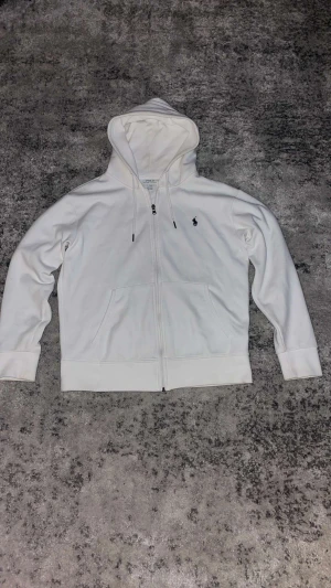 Vit hoodie från Polo Ralph Lauren - Vit hoodie från Polo Ralph Lauren med dragkedja och huva. Tröjan har två fickor framtill och den klassiska Polo-loggan broderad på bröstet. Långärmad och tillverkad i mjuk bomull, perfekt för chill dagar. Snygg och enkel design som funkar året runt. Säljer denna eftersom att den är för liten 
