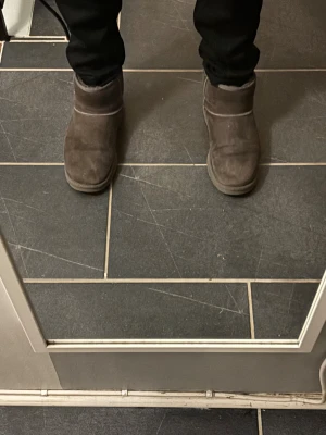 Bruna UGG boots i mocka - Mysiga bruna UGG boots i klassisk modell med rund tå och platt sula. Skorna är fodrade med mjuk päls på insidan och har en ovandel i mocka. Perfekta för kalla dagar och ger en chill vibe till din outfit.