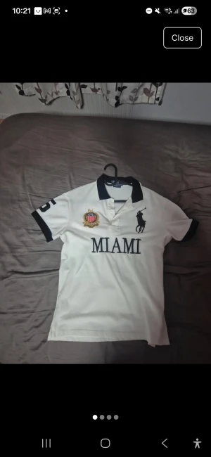 Vit Polo Ralph Lauren Miami piké - Vit pikétröja från Polo Ralph Lauren med svart krage och svarta detaljer på ärmarna. Stor broderad logga på bröstet, Miami-tryck och patch med emblem. Siffra 5 på ena ärmen. Custom fit-modell i bomull, perfekt för dig som gillar sportig och clean stil.