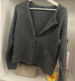 Mörkgrå kofta med knappar - Säljer en mörkgrå kofta från H&M med mjukt stickat material och klassisk v-ringning. Koftan har långa ärmar och stängs med fyra knappar framtill. Perfekt för lager-på-lager och chill stil.