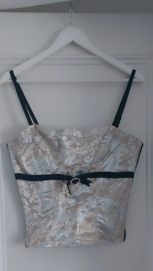 Silvrig blommig korsett med rosett - Snygg korsett-topp med blommigt mönster i silver och beige, svarta axelband och svart band med liten rosett och strassdetalj framtill. Toppen har croppad passform och är tillverkad i glansigt satinliknande material. Perfekt för dig som vill sticka ut med en elegant vibe.