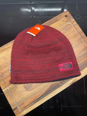 Röd stickad mössa från The North Face - Snygg röd och svart stickad mössa från The North Face med broderad logga framtill. Mössan har ett mjukt fleecefoder på insidan för extra värme och komfort. Perfekt för kalla dagar och outdoor-aktiviteter. Klassisk passform och diskret etikett i sidan. Oanvänd 