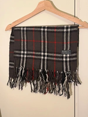 Burberry halsduk - Snygg rutig halsduk från Burberrys i 100% lammull, tillverkad i England. Färgerna är mörkgrå, svart, vit och röd med klassiskt rutmönster och långa fransar i ändarna. Perfekt för att ge din outfit en cool och tidlös vibe. Självklart äkta!