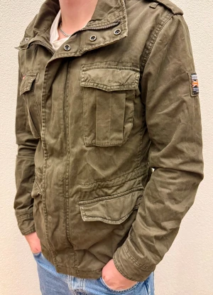 Olivgrön fieldjacket från Superdry - Säljer en olivgrön fieldjacket från Superdry med coola militärvibbar. Jackan har flera fickor med lock, hög krage med metallöljetter och patch på ärmen. 