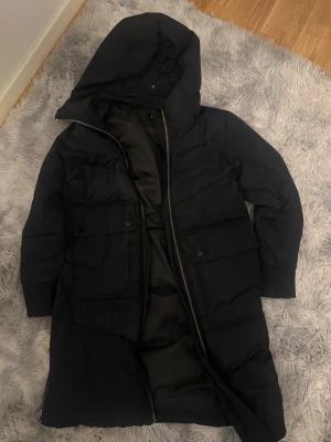 Svart lång pufferjacka Everest - Svart lång pufferjacka från Everest med huva, stora fickor med knappar och dragkedja framtill. Jackan har även dragkedjor i nederkant på baksidan och ribbade muddar vid ärmslut. Perfekt för kalla dagar och har en clean, stilren look.