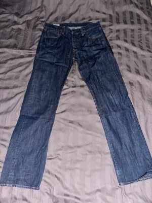 Levi's 501 blå jeans straight fit STORLEK: 30/32 - Klassiska Levi's 501 jeans i mörkblå tvätt med raka ben och fem fickor. Jeansen har knappgylf och orangea sömmar som ger en snygg kontrast. Perfekt för dig som gillar tidlös stil och vill ha ett par jeans som funkar till allt.